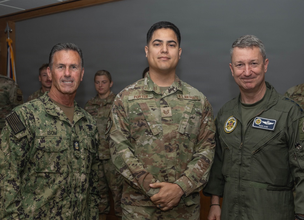 Three military personnel pose together for a formal photo in what appears to be an indoor military facility. The individuals represent different branches of the U.S. Armed Forces, including the U.S. Navy and U.S. Air Force. Additional uniformed personnel are visible in the background.