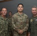 Three military personnel pose together for a formal photo in what appears to be an indoor military facility. The individuals represent different branches of the U.S. Armed Forces, including the U.S. Navy and U.S. Air Force. Additional uniformed personnel are visible in the background.
