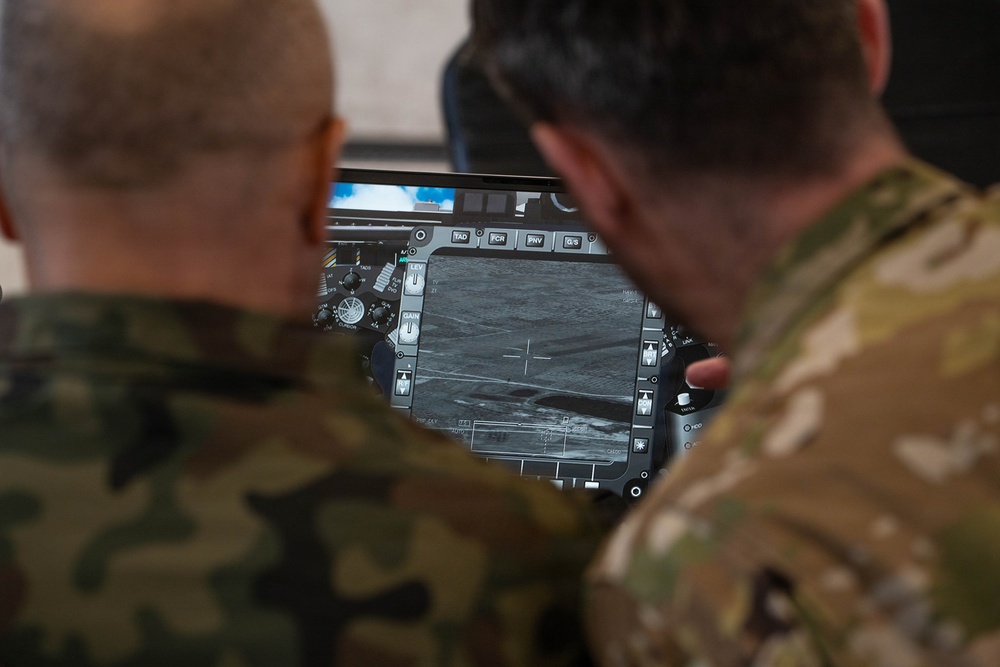 Two military personnel in different camouflage uniforms are viewing a tactical display screen showing what appears to be an infrared or thermal imaging feed of terrain. The screen interface includes multiple control buttons and overlays consistent with a drone ground control station or targeting system. One individual appears to be pointing at or operating the display.