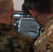 Two military personnel in different camouflage uniforms are viewing a tactical display screen showing what appears to be an infrared or thermal imaging feed of terrain. The screen interface includes multiple control buttons and overlays consistent with a drone ground control station or targeting system. One individual appears to be pointing at or operating the display.