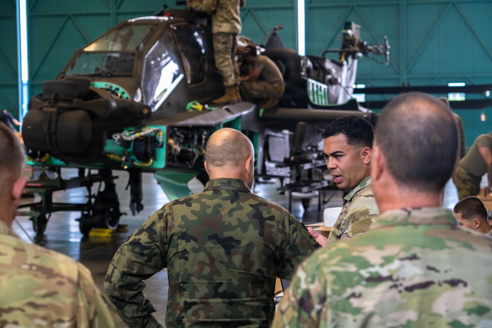 Military personnel in a hangar are gathered around a partially disassembled attack helicopter, likely an AH-64 Apache, engaged in what appears to be a maintenance briefing or training exercise. Soldiers wearing different camouflage patterns — including US Army OCP and what appears to be a European woodland pattern — are conversing, suggesting a multinational or joint military cooperation context. One soldier atop the helicopter is actively performing maintenance work.