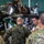 Polish General Piotr Trytek Strengthens Alliance During 12th CAB Visit Military personnel in a hangar are gathered around a partially disassembled attack helicopter, likely an AH-64 Apache, engaged in what appears to be a maintenance briefing or training exercise. Soldiers wearing different camouflage patterns — including US Army OCP and what appears to be a European woodland pattern — are conversing, suggesting a multinational or joint military cooperation context. One soldier atop the helicopter is actively performing maintenance work.