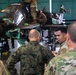 Military personnel in a hangar are gathered around a partially disassembled attack helicopter, likely an AH-64 Apache, engaged in what appears to be a maintenance briefing or training exercise. Soldiers wearing different camouflage patterns — including US Army OCP and what appears to be a European woodland pattern — are conversing, suggesting a multinational or joint military cooperation context. One soldier atop the helicopter is actively performing maintenance work.