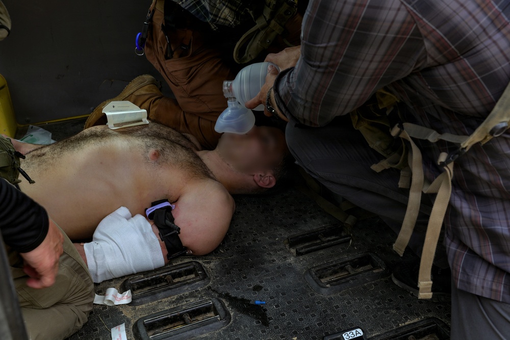 A shirtless male casualty is lying face-down on what appears to be the floor of a military vehicle or aircraft, receiving emergency medical treatment. At least two individuals are administering care, including bag-valve-mask (BVM) ventilation and wound bandaging. The scene indicates a tactical combat casualty care (TCCC) scenario.