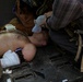 A shirtless male casualty is lying face-down on what appears to be the floor of a military vehicle or aircraft, receiving emergency medical treatment. At least two individuals are administering care, including bag-valve-mask (BVM) ventilation and wound bandaging. The scene indicates a tactical combat casualty care (TCCC) scenario.