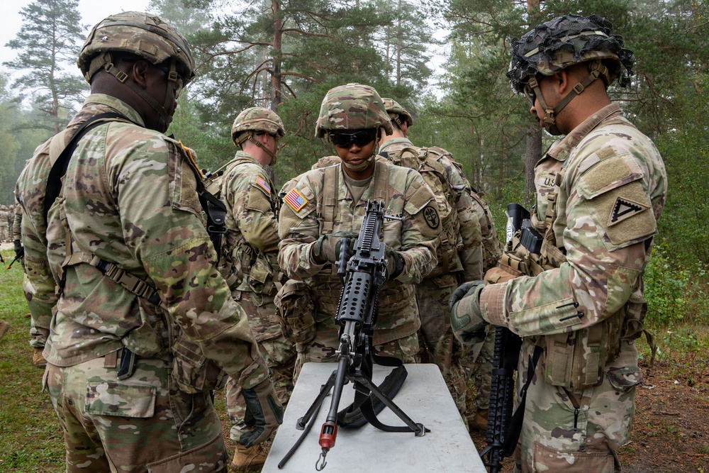 A group of U.S. Army soldiers in Operational Camouflage Pattern (OCP) uniforms are gathered around a field table during what appears to be a weapons training or maintenance exercise. A central soldier is handling and inspecting what appears to be an M249 Squad Automatic Weapon (SAW) or similar light machine gun on the table. The setting is an outdoor wooded training area under overcast conditions.