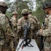 A group of U.S. Army soldiers in Operational Camouflage Pattern (OCP) uniforms are gathered around a field table during what appears to be a weapons training or maintenance exercise. A central soldier is handling and inspecting what appears to be an M249 Squad Automatic Weapon (SAW) or similar light machine gun on the table. The setting is an outdoor wooded training area under overcast conditions.