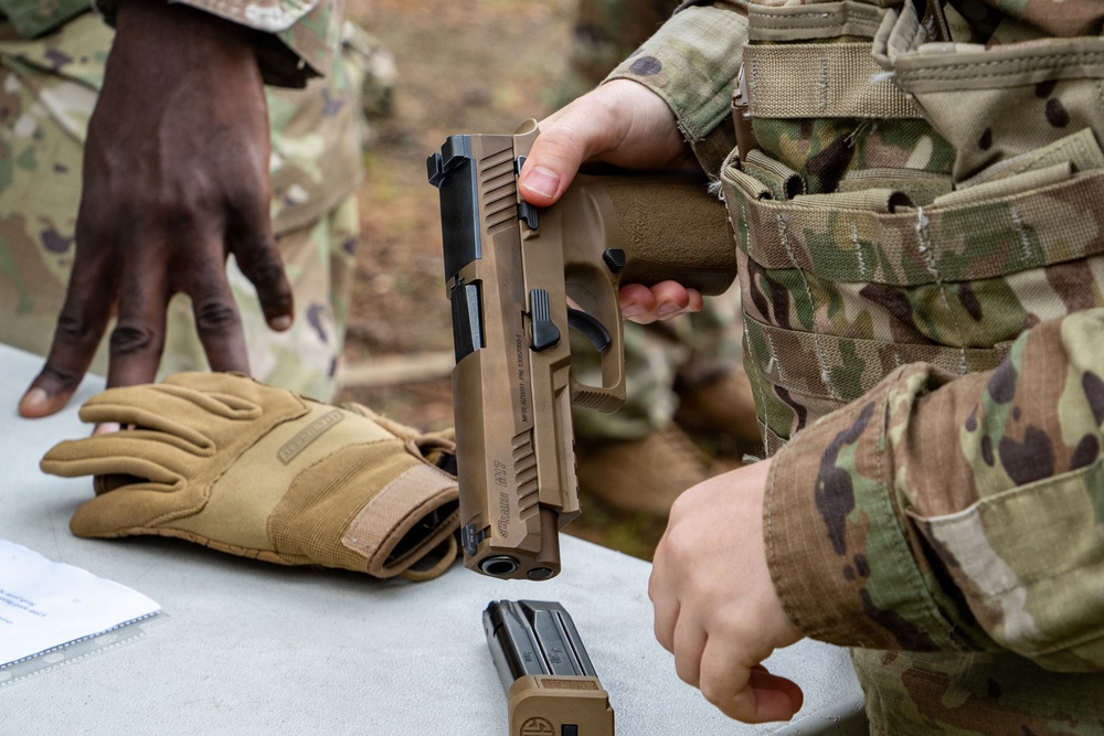 Military personnel in OCP (Operational Camouflage Pattern) uniforms are handling a SIG Sauer M17/M18 pistol at what appears to be a training or qualification range. One soldier is holding the pistol with the slide locked back, indicating a cleared or unloaded weapon, while a detached magazine sits on the table. Tan tactical gloves and paperwork are also visible on the surface.