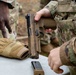 Military personnel in OCP (Operational Camouflage Pattern) uniforms are handling a SIG Sauer M17/M18 pistol at what appears to be a training or qualification range. One soldier is holding the pistol with the slide locked back, indicating a cleared or unloaded weapon, while a detached magazine sits on the table. Tan tactical gloves and paperwork are also visible on the surface.