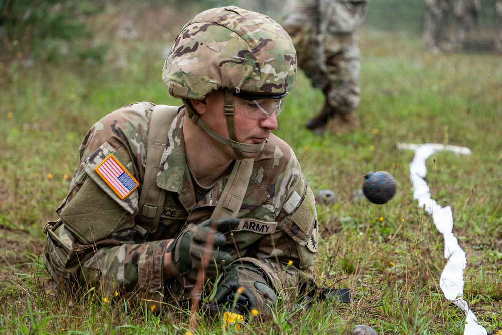 A U.S. Army soldier in MultiCam (OCP) camouflage uniform is prone on the ground, closely examining what appears to be simulated mines or explosive devices marked with white tape/cord during a training exercise. The soldier is wearing full combat gear including a helmet, protective eyewear, and tactical gloves. A second soldier is visible in the blurred background.