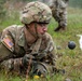 A U.S. Army soldier in MultiCam (OCP) camouflage uniform is prone on the ground, closely examining what appears to be simulated mines or explosive devices marked with white tape/cord during a training exercise. The soldier is wearing full combat gear including a helmet, protective eyewear, and tactical gloves. A second soldier is visible in the blurred background.