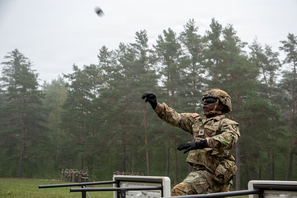 A U.S. Army soldier in full combat gear is throwing what appears to be a grenade during a training exercise at an outdoor range. The soldier is positioned behind a barrier structure, wearing OCP (Operational Camouflage Pattern) uniform, helmet, protective eyewear, and tactical gloves. A group of soldiers observing the exercise is visible in the background near a treeline.