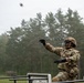 A U.S. Army soldier in full combat gear is throwing what appears to be a grenade during a training exercise at an outdoor range. The soldier is positioned behind a barrier structure, wearing OCP (Operational Camouflage Pattern) uniform, helmet, protective eyewear, and tactical gloves. A group of soldiers observing the exercise is visible in the background near a treeline.
