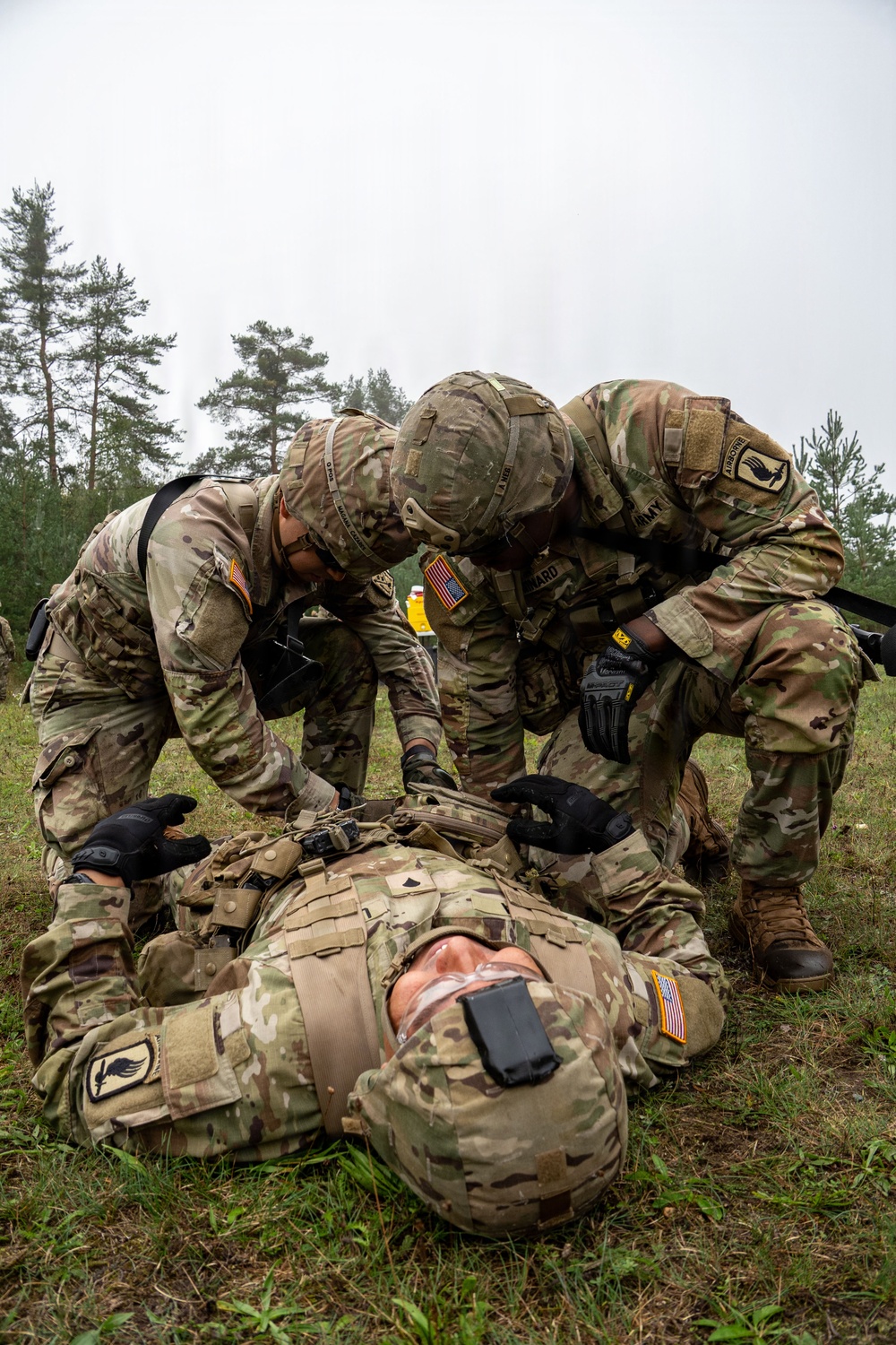 Two U.S. Army soldiers in MultiCam/OCP uniforms are performing tactical combat casualty care (TCCC) on a third soldier lying on the ground. All personnel wear U.S. flag patches and unit patches consistent with an Airborne division. The setting appears to be an outdoor field training exercise in a forested area under overcast skies.