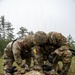 Two U.S. Army soldiers in MultiCam/OCP uniforms are performing tactical combat casualty care (TCCC) on a third soldier lying on the ground. All personnel wear U.S. flag patches and unit patches consistent with an Airborne division. The setting appears to be an outdoor field training exercise in a forested area under overcast skies.