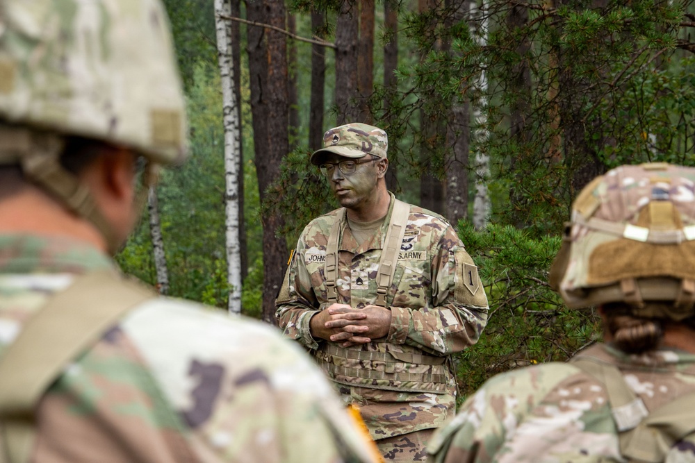 A U.S. Army soldier, partially identified by the name tag 'JOHN-' and 'U.S. ARMY' tape, addresses a group of fellow soldiers in a forested training environment. The central figure wears camouflage face paint, a patrol cap, glasses, and OCP (Operational Camouflage Pattern) uniform with a tactical vest. The setting appears to be an outdoor field training exercise or briefing.
