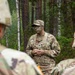 A U.S. Army soldier, partially identified by the name tag 'JOHN-' and 'U.S. ARMY' tape, addresses a group of fellow soldiers in a forested training environment. The central figure wears camouflage face paint, a patrol cap, glasses, and OCP (Operational Camouflage Pattern) uniform with a tactical vest. The setting appears to be an outdoor field training exercise or briefing.