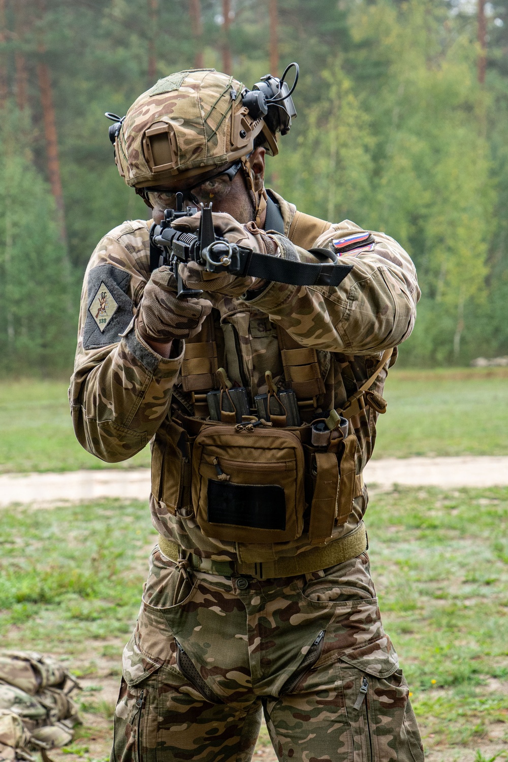 A soldier in full tactical gear is in a shooting stance, aiming a rifle with a sling attached. The individual is wearing multicam camouflage uniform, a tactical helmet with night-vision mount, and a plate carrier vest with multiple magazine pouches. A flag patch visible on the right shoulder appears to be Slovenian.