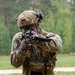 A soldier in full tactical gear is in a shooting stance, aiming a rifle with a sling attached. The individual is wearing multicam camouflage uniform, a tactical helmet with night-vision mount, and a plate carrier vest with multiple magazine pouches. A flag patch visible on the right shoulder appears to be Slovenian.