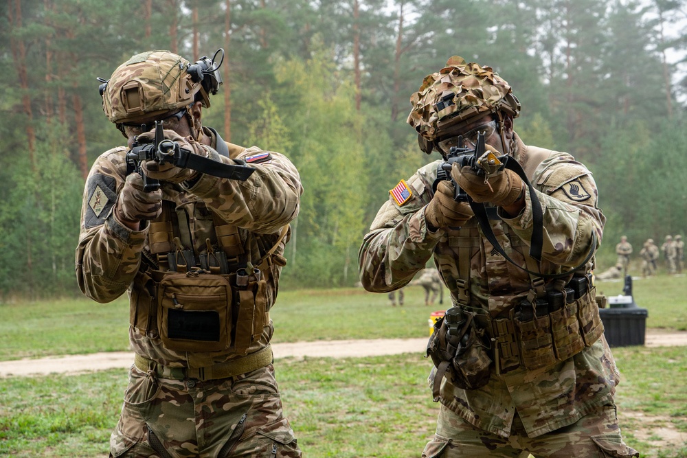 Two soldiers in MultiCam camouflage uniforms are aiming rifles in what appears to be a training exercise at an outdoor range. Both wear tactical gear including plate carriers, helmets, and gloves, with US Army patches visible on the right soldier. Additional personnel are visible in the background near a forested area.