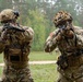 Two soldiers in MultiCam camouflage uniforms are aiming rifles in what appears to be a training exercise at an outdoor range. Both wear tactical gear including plate carriers, helmets, and gloves, with US Army patches visible on the right soldier. Additional personnel are visible in the background near a forested area.