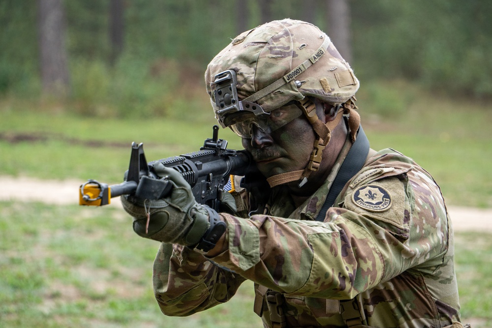 A U.S. Army soldier in full combat gear is aiming a rifle in an outdoor training environment. The soldier is wearing MultiCam (OCP) camouflage uniform, a tactical helmet with accessories, and has face paint applied. Visible unit patches and helmet markings suggest an active-duty or National Guard assignment.