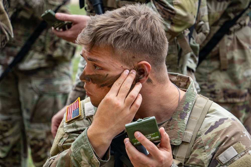 A U.S. Army soldier in Operational Camouflage Pattern (OCP) uniform is applying camouflage face paint using a compact mirror. The soldier is holding a green military-issue face paint compact and applying dark paint near their temple and cheek area. Other soldiers in similar uniforms are visible in the background.