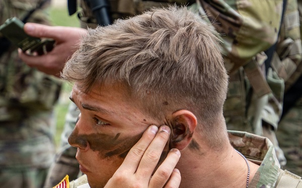 A U.S. Army soldier in Operational Camouflage Pattern (OCP) uniform is applying camouflage face paint using a compact mirror. The soldier is holding a green military-issue face paint compact and applying dark paint near their temple and cheek area. Other soldiers in similar uniforms are visible in the background.