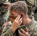 A U.S. Army soldier in Operational Camouflage Pattern (OCP) uniform is applying camouflage face paint using a compact mirror. The soldier is holding a green military-issue face paint compact and applying dark paint near their temple and cheek area. Other soldiers in similar uniforms are visible in the background.