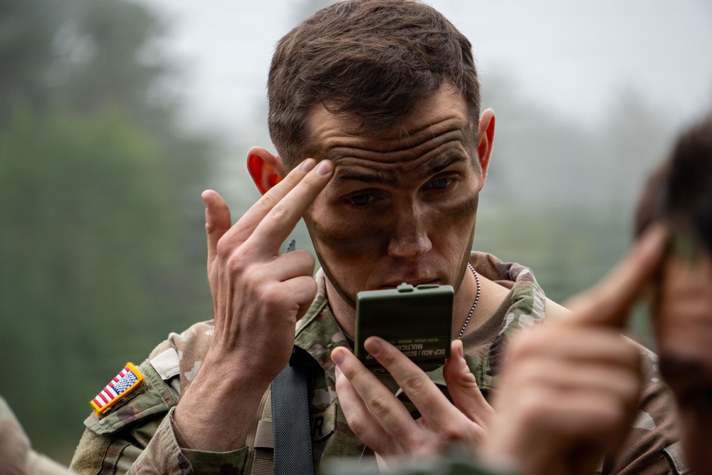 A U.S. Army soldier in Operational Camouflage Pattern (OCP) uniform is applying face camouflage paint using a small military-issue compact mirror. The soldier is using his fingers to apply or spread the paint on his forehead area. The setting appears to be an outdoor field environment with overcast, foggy conditions.