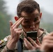 A U.S. Army soldier in Operational Camouflage Pattern (OCP) uniform is applying face camouflage paint using a small military-issue compact mirror. The soldier is using his fingers to apply or spread the paint on his forehead area. The setting appears to be an outdoor field environment with overcast, foggy conditions.