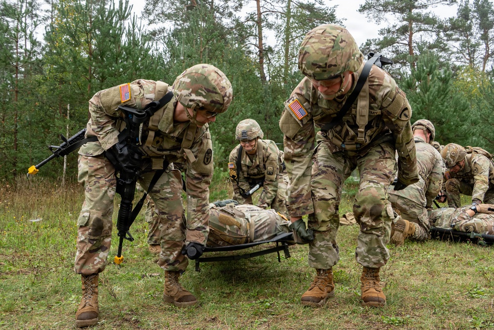 U.S. Army soldiers in full combat gear are conducting a field medical casualty evacuation (CASEVAC) training exercise in a wooded outdoor environment. Two soldiers in the foreground are lifting a simulated casualty on a litter/stretcher, while additional soldiers in the background attend to other simulated casualties on the ground. The presence of training weapons with yellow safety markers and the controlled setting indicate this is a military training exercise rather than a combat situation.