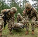 U.S. Army soldiers in full combat gear are conducting a field medical casualty evacuation (CASEVAC) training exercise in a wooded outdoor environment. Two soldiers in the foreground are lifting a simulated casualty on a litter/stretcher, while additional soldiers in the background attend to other simulated casualties on the ground. The presence of training weapons with yellow safety markers and the controlled setting indicate this is a military training exercise rather than a combat situation.