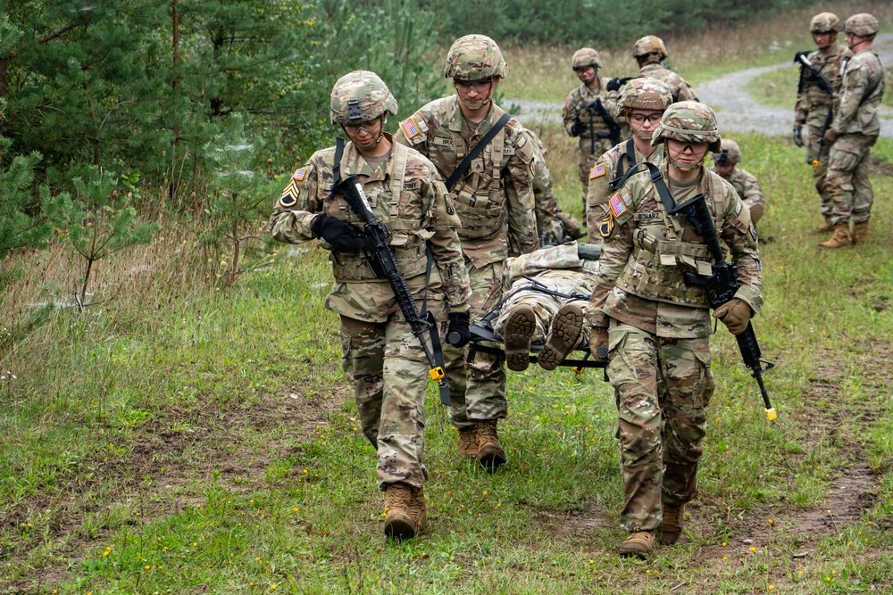 U.S. Army soldiers in OCP (Operational Camouflage Pattern) uniforms are conducting a casualty evacuation (CASEVAC) training exercise in a wooded field environment. Three soldiers in the foreground are carrying a simulated casualty on a litter/stretcher, while additional soldiers are visible in the background. The scene appears to be a field training exercise (FTX) based on the organized nature of the activity and presence of multiple personnel.