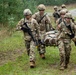 U.S. Army soldiers in OCP (Operational Camouflage Pattern) uniforms are conducting a casualty evacuation (CASEVAC) training exercise in a wooded field environment. Three soldiers in the foreground are carrying a simulated casualty on a litter/stretcher, while additional soldiers are visible in the background. The scene appears to be a field training exercise (FTX) based on the organized nature of the activity and presence of multiple personnel.