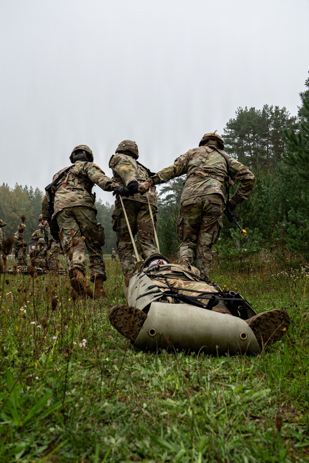 Three soldiers in camouflage uniforms and helmets are dragging a casualty on an inflatable litter/sled across a grassy field during what appears to be a military training exercise. The casualty is strapped onto an olive-drab inflatable rescue sled and is being pulled by ropes held by the soldiers. The overcast sky and forested background suggest a temperate environment, likely a training area in Europe.
