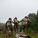Three soldiers in camouflage uniforms and helmets are dragging a casualty on an inflatable litter/sled across a grassy field during what appears to be a military training exercise. The casualty is strapped onto an olive-drab inflatable rescue sled and is being pulled by ropes held by the soldiers. The overcast sky and forested background suggest a temperate environment, likely a training area in Europe.