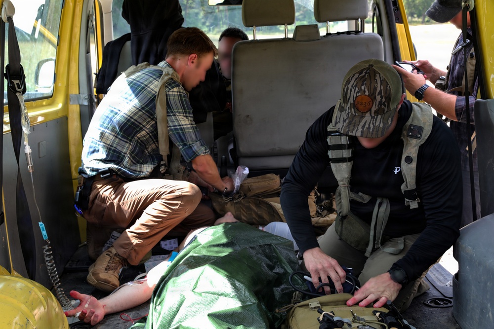 The image shows what appears to be a tactical medical response or casualty care scenario taking place inside a yellow van or vehicle. Two individuals wearing tactical vests are providing care to a person lying on the floor covered with a green tarp, with visible medical supplies and equipment present. A third person in the background appears to be monitoring or documenting the situation with a phone.