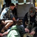 The image shows what appears to be a tactical medical response or casualty care scenario taking place inside a yellow van or vehicle. Two individuals wearing tactical vests are providing care to a person lying on the floor covered with a green tarp, with visible medical supplies and equipment present. A third person in the background appears to be monitoring or documenting the situation with a phone.