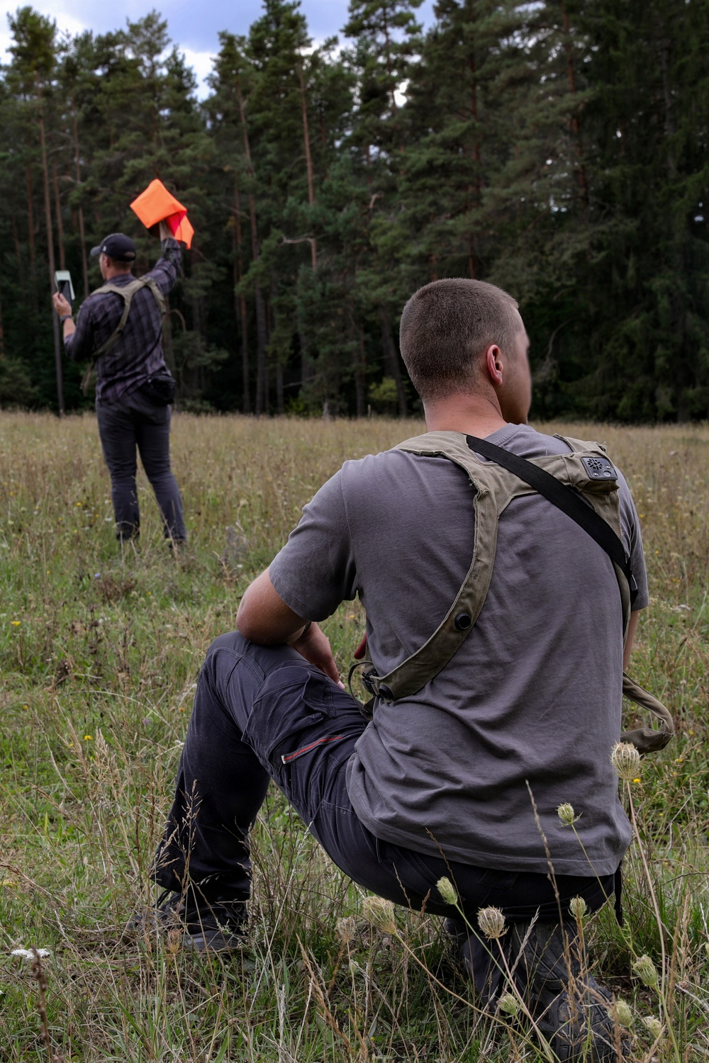 Two individuals are in an open grassy field bordered by a dense pine forest. One person is seated in the foreground wearing a gray t-shirt and olive sling bag, while the other stands in the background holding an orange signal flag raised above their head and holding a device in the other hand. The scene appears to involve signaling or surveying activity in an outdoor setting.