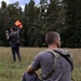 Two individuals are in an open grassy field bordered by a dense pine forest. One person is seated in the foreground wearing a gray t-shirt and olive sling bag, while the other stands in the background holding an orange signal flag raised above their head and holding a device in the other hand. The scene appears to involve signaling or surveying activity in an outdoor setting.
