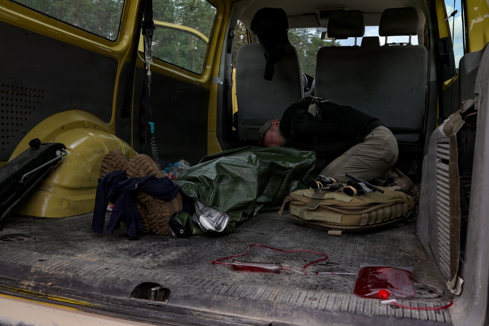 The image shows the interior cargo area of a yellow van being used as a field medical evacuation vehicle. A person in dark tactical clothing is leaning over what appears to be a casualty wrapped in a green tarp or emergency blanket. Blood and medical tubing are visible on the floor of the vehicle, indicating active or recent medical treatment.
