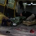 The image shows the interior cargo area of a yellow van being used as a field medical evacuation vehicle. A person in dark tactical clothing is leaning over what appears to be a casualty wrapped in a green tarp or emergency blanket. Blood and medical tubing are visible on the floor of the vehicle, indicating active or recent medical treatment.