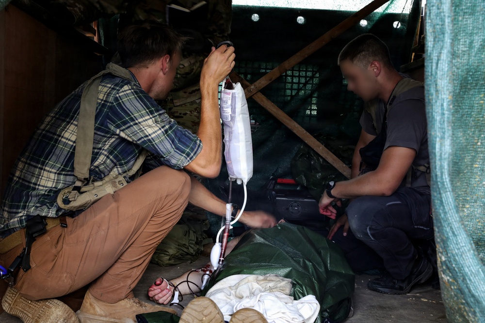 Two individuals are providing emergency medical care to a casualty lying on the ground inside a makeshift shelter or fortified position. One person is administering an IV drip while the other attends to the patient from the opposite side. The setting appears to be a field medical treatment point in a conflict or military training environment.