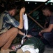 Two individuals are providing emergency medical care to a casualty lying on the ground inside a makeshift shelter or fortified position. One person is administering an IV drip while the other attends to the patient from the opposite side. The setting appears to be a field medical treatment point in a conflict or military training environment.