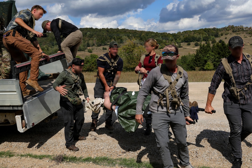 A group of approximately seven individuals in tactical/mixed civilian clothing are transferring a person on a stretcher or litter from the bed of a utility vehicle onto the ground. The scene appears to take place on a gravel road in a rural, hilly environment. This appears consistent with a medical evacuation drill or tactical casualty care training exercise.