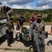 A group of approximately seven individuals in tactical/mixed civilian clothing are transferring a person on a stretcher or litter from the bed of a utility vehicle onto the ground. The scene appears to take place on a gravel road in a rural, hilly environment. This appears consistent with a medical evacuation drill or tactical casualty care training exercise.