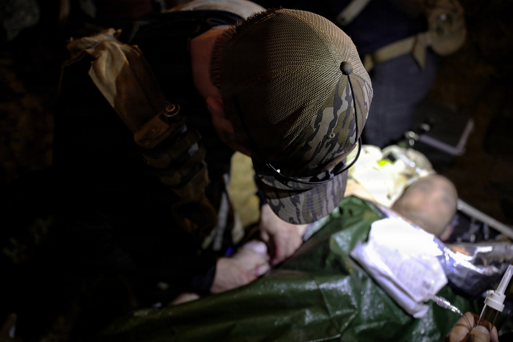 A person wearing camouflage clothing and a camouflage cap is bent over, performing what appears to be a medical procedure or treatment on a patient lying on a green surface. The scene is dimly lit, with medical supplies visible nearby including what appears to be an IV bag and syringe. The setting suggests a field medical or tactical medical scenario.
