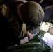 A person wearing camouflage clothing and a camouflage cap is bent over, performing what appears to be a medical procedure or treatment on a patient lying on a green surface. The scene is dimly lit, with medical supplies visible nearby including what appears to be an IV bag and syringe. The setting suggests a field medical or tactical medical scenario.