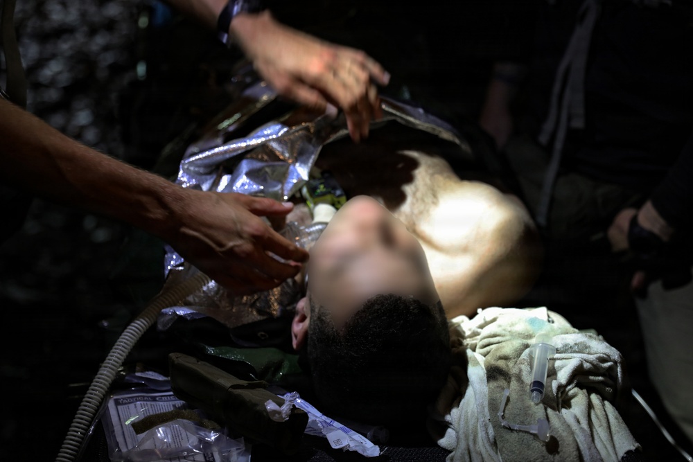 A person is receiving emergency medical treatment in a low-light environment. Medical personnel are attending to the patient, who is lying supine and appears to have an airway management device in place. Various medical supplies are visible around the patient.